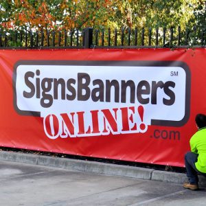 A red banner advertising SignsBanners ONLINE! on a fence outdoors.